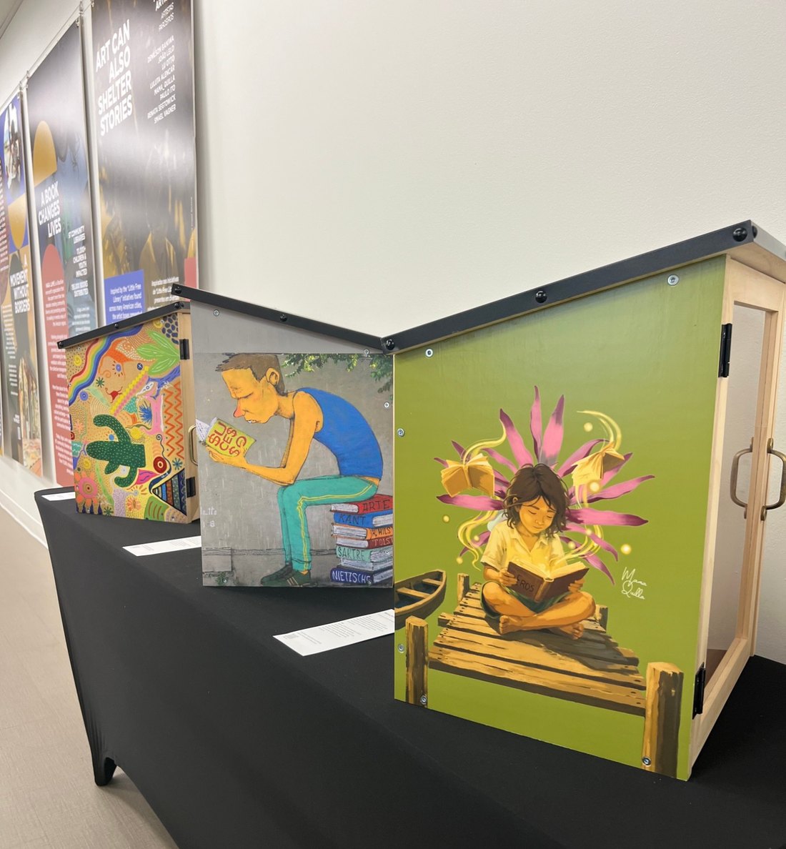Três bibliotecas de madeira pintadas sobre um pedestal, com os banners da exposição na parede atrás